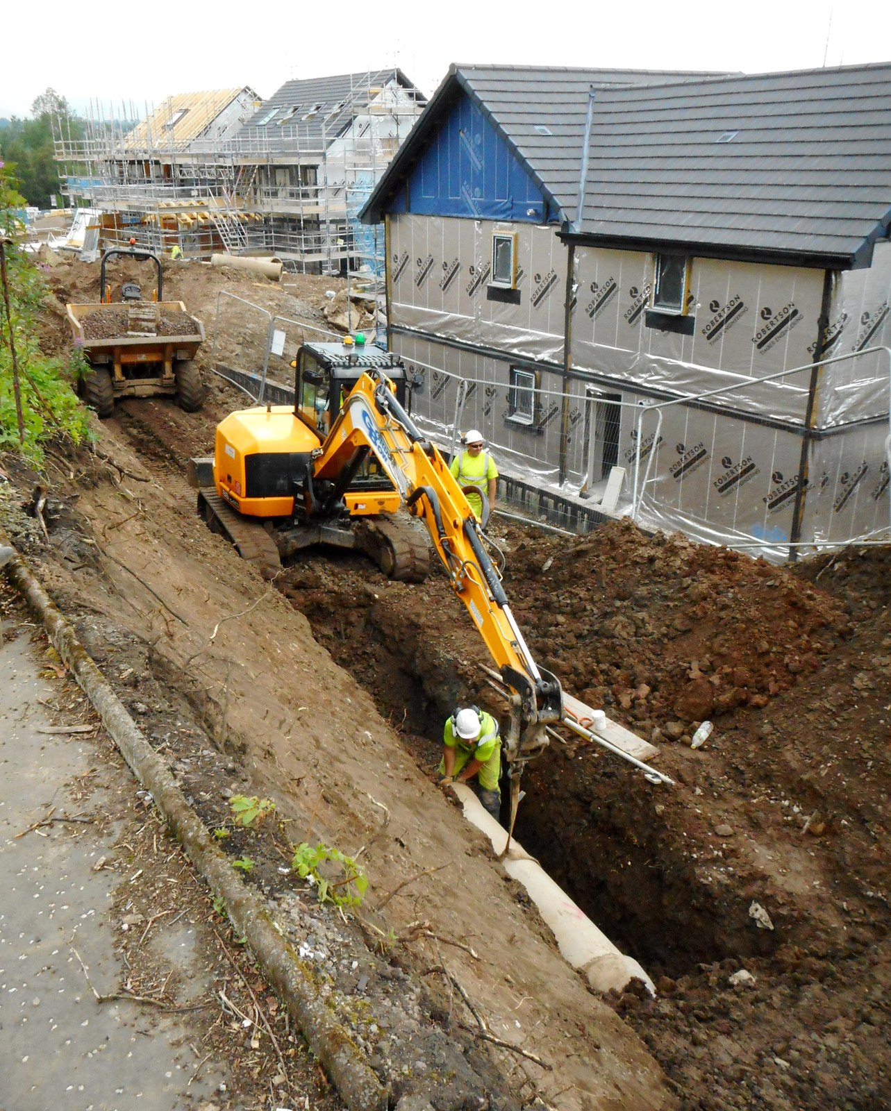 Excavator laying drainage in an open trench beside new housing